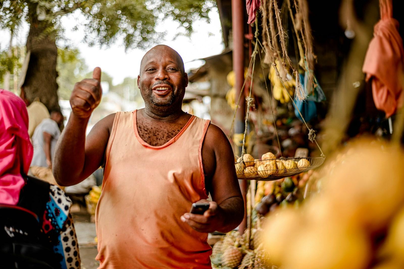 A shop owner in Dar es Salaam giving a thumbs up while holding his phone, standing in front of his fruit stand